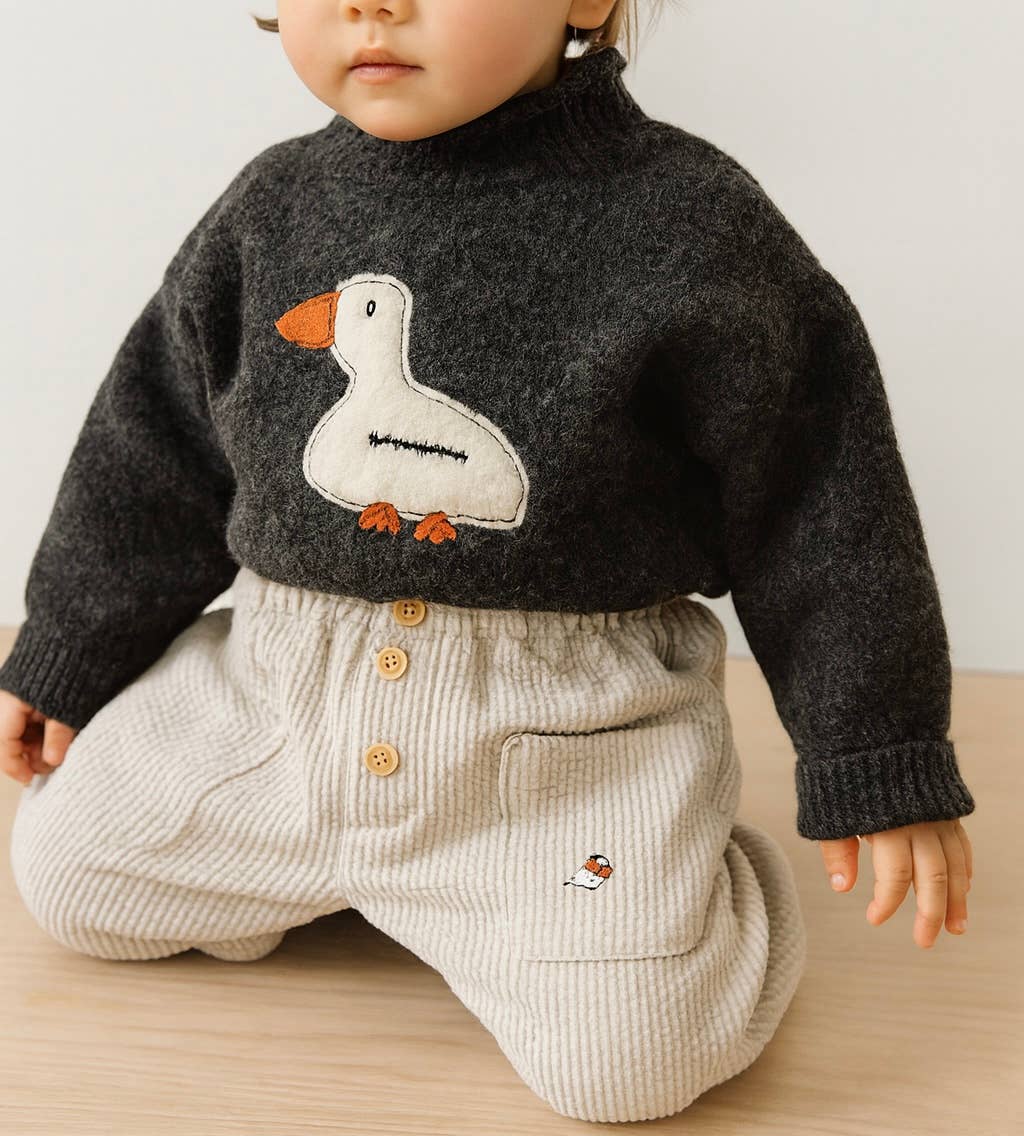 Child wearing a dark sweater with a duck design and light-colored pants on a wooden surface.