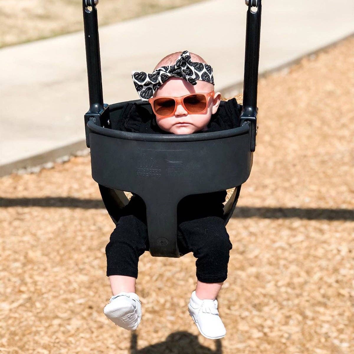 Baby in a swing wearing sunglasses and a headband on a playground.