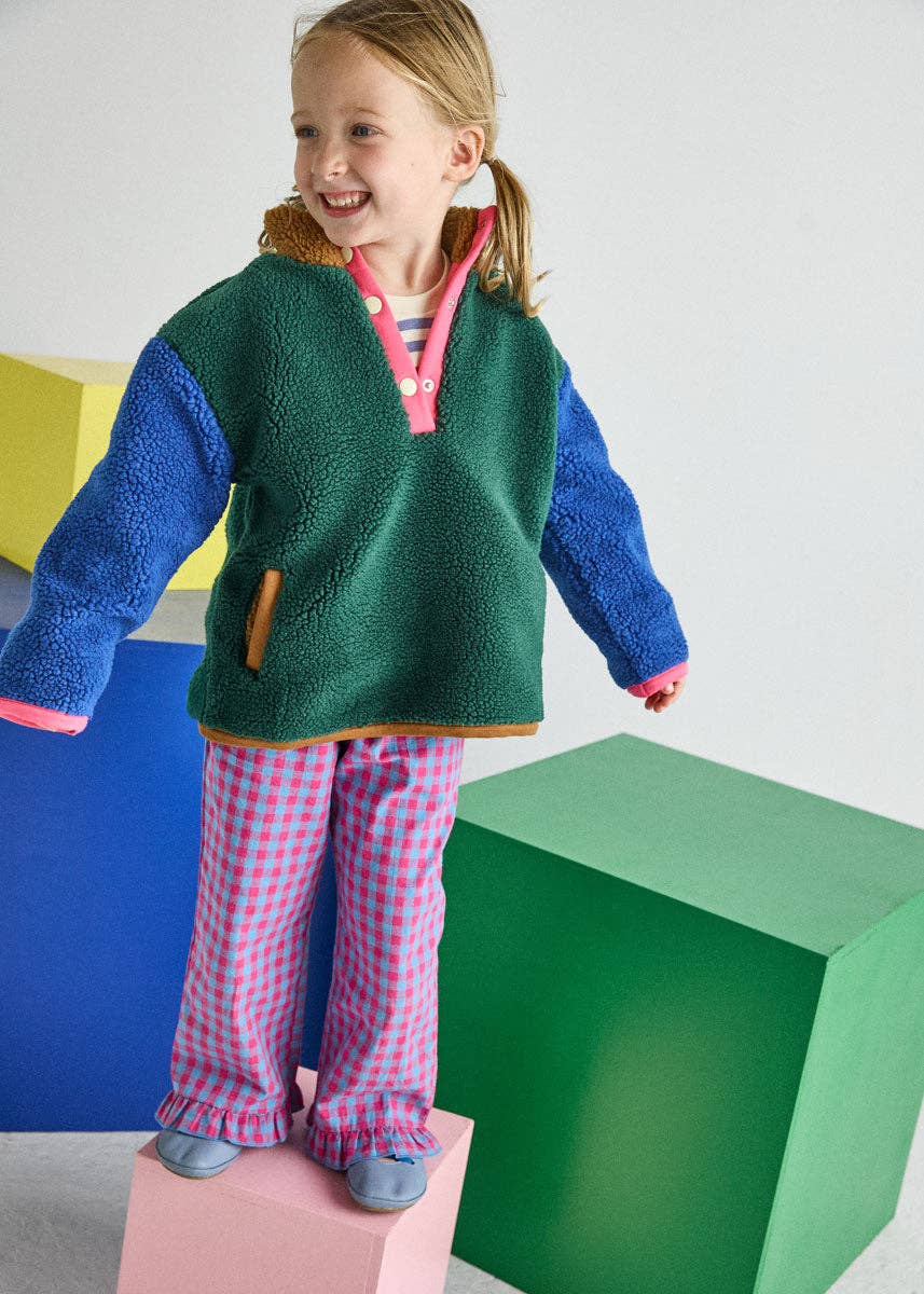 A young girl stands on a pink block wearing a green and blue fleece pullover with pink trim and pink-and-white gingham pajama pants, in a brightly lit studio with colorful geometric blocks.