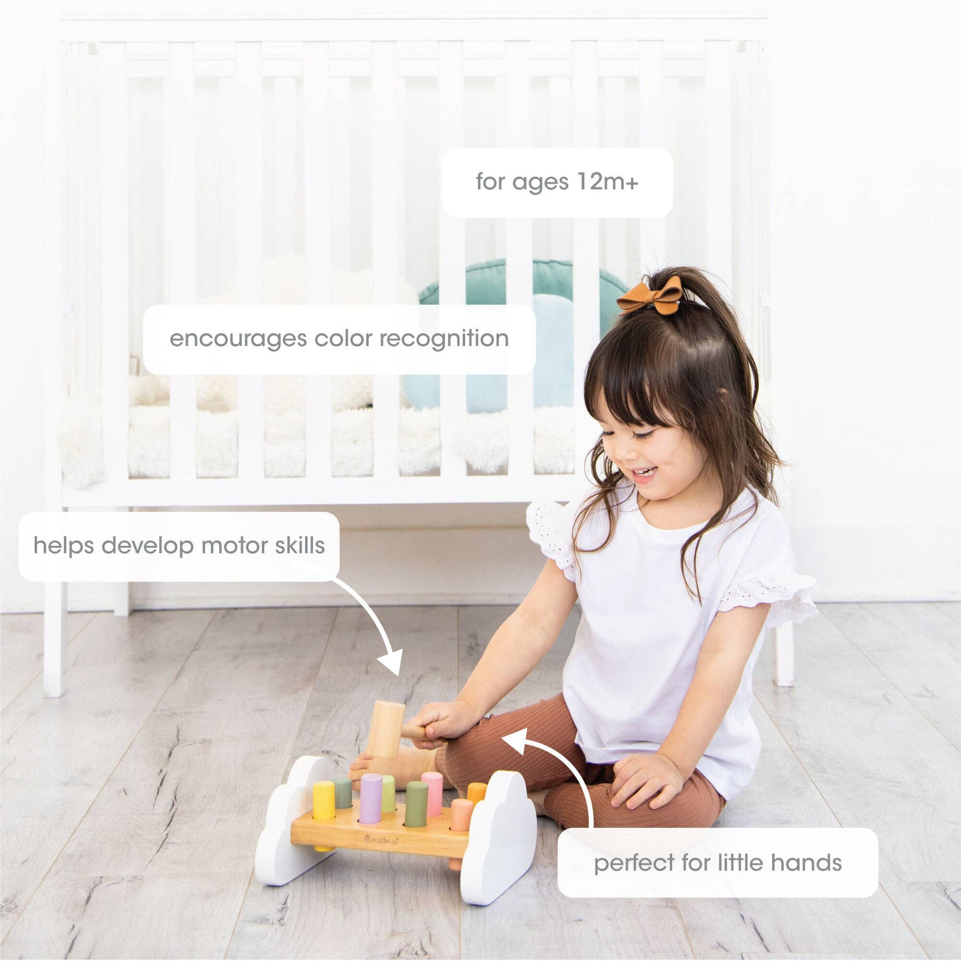 A young child sits on a wooden floor in front of a crib playing with a wooden hammer bench toy with colorful pegs and a wooden mallet.