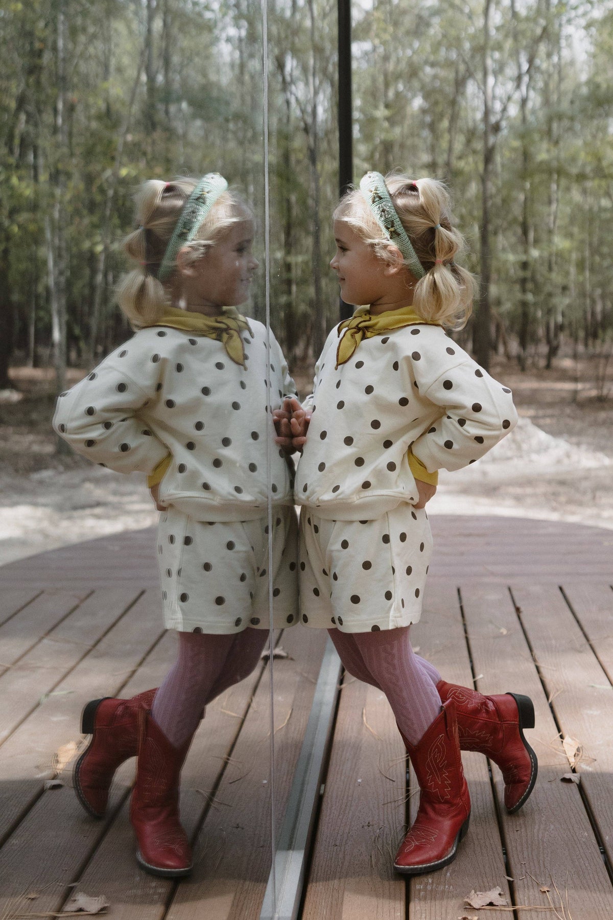 A young girl stands on a wooden deck in front of a mirror, wearing a cream sweatshirt and shorts with brown polka dots, a yellow neck scarf, pink tights, and red cowboy boots. Her reflection is visible in the mirror, with a forest background.