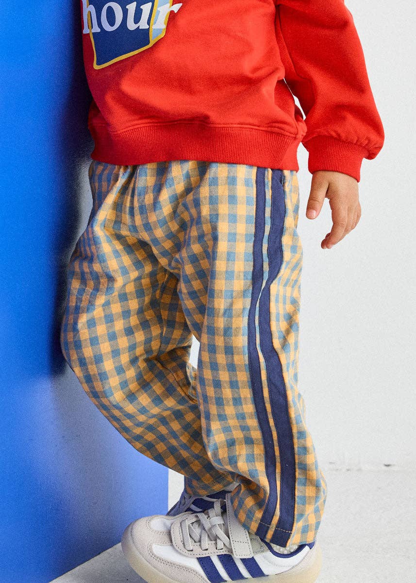 A child wearing yellow and blue gingham check pants with navy blue side stripes, paired with a red sweatshirt and white sneakers, leaning against a blue wall.