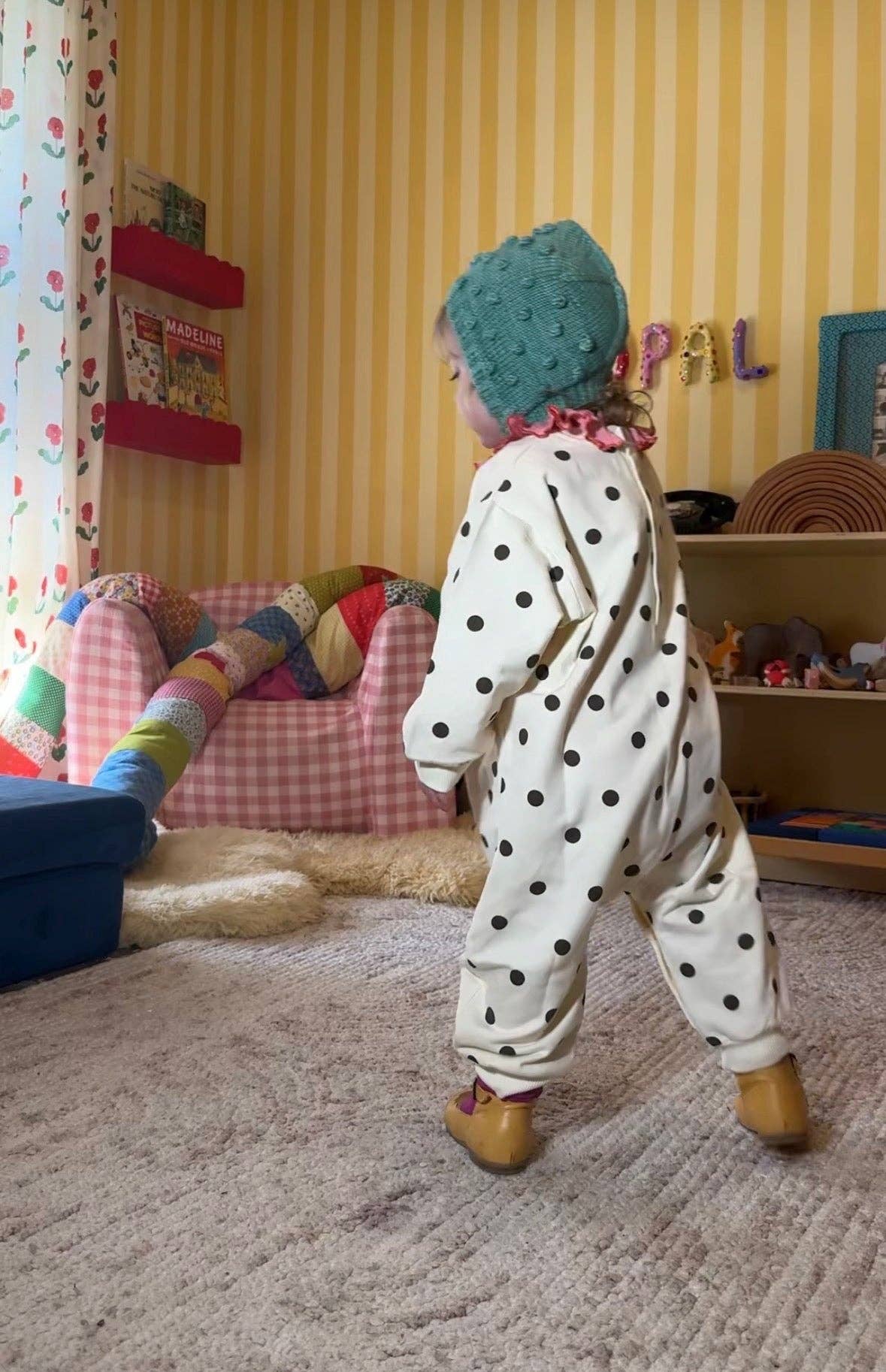 A toddler standing in a playroom, wearing a cream-colored one-piece jumpsuit with brown polka dots, a teal knit hat, and tan boots.