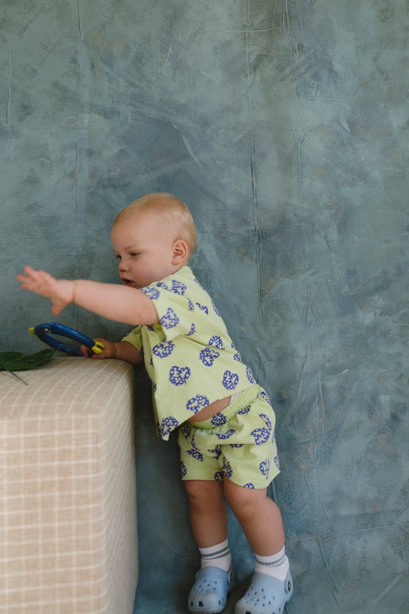 Baby in a pretzel outfit standing against a textured blue wall.