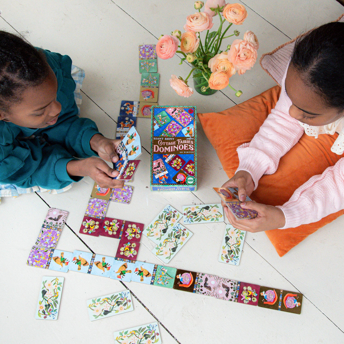 Two children playing with dominoes on a tiled floor with a vase of flowers in the background.
