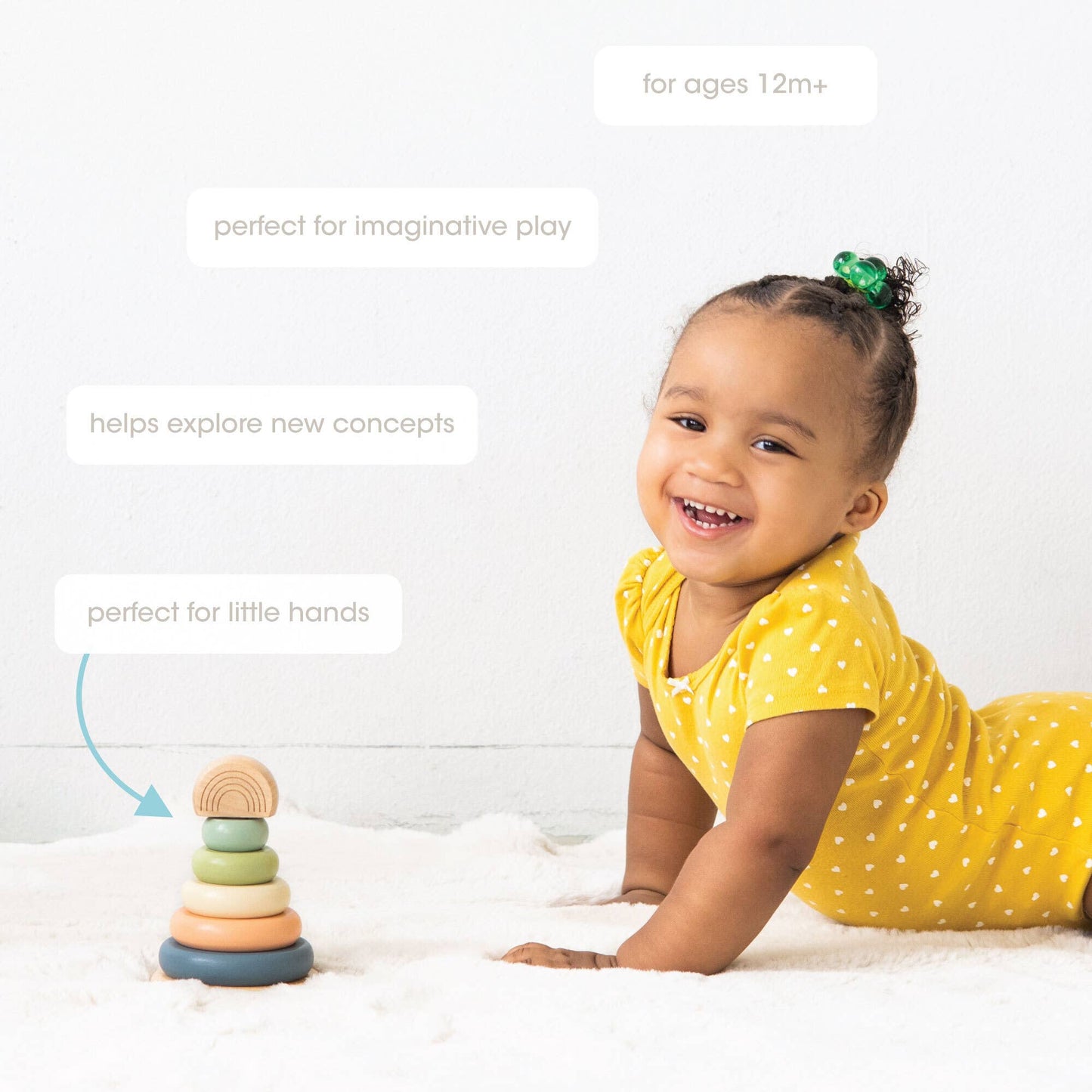 A young child wearing a yellow polka dot shirt smiling next to a wooden stacking toy consisting of a peg with five multicolor earth tone rings and a wooden rainbow topper on a white background.