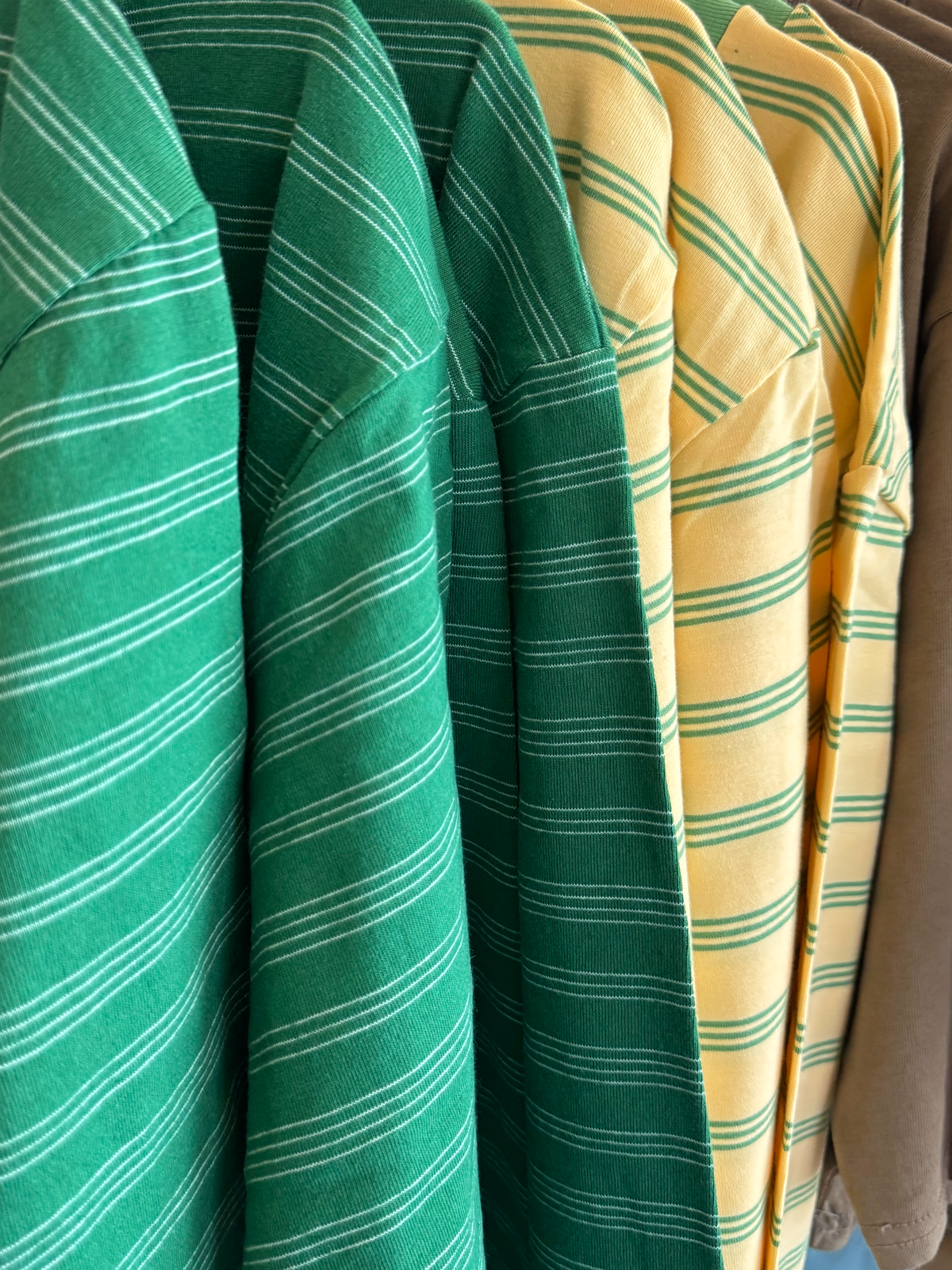 A close-up of striped collared shirts hanging on a rack, featuring a green shirt with thin white stripes, a green shirt with darker green stripes, and a yellow shirt with green stripes.