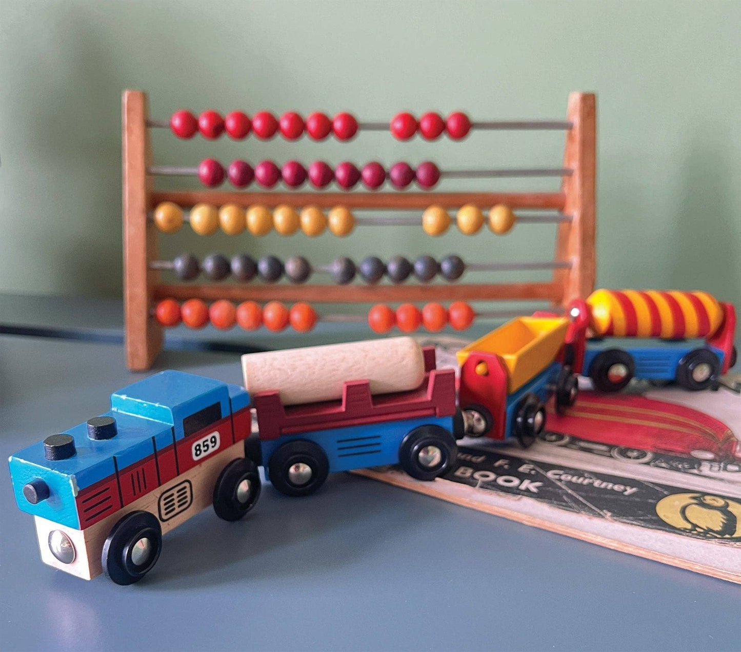 A wooden train set with one engine and three colorful attached carriages designed to resemble industrial cargo goods, displayed on a table top with an abacus toy in the background