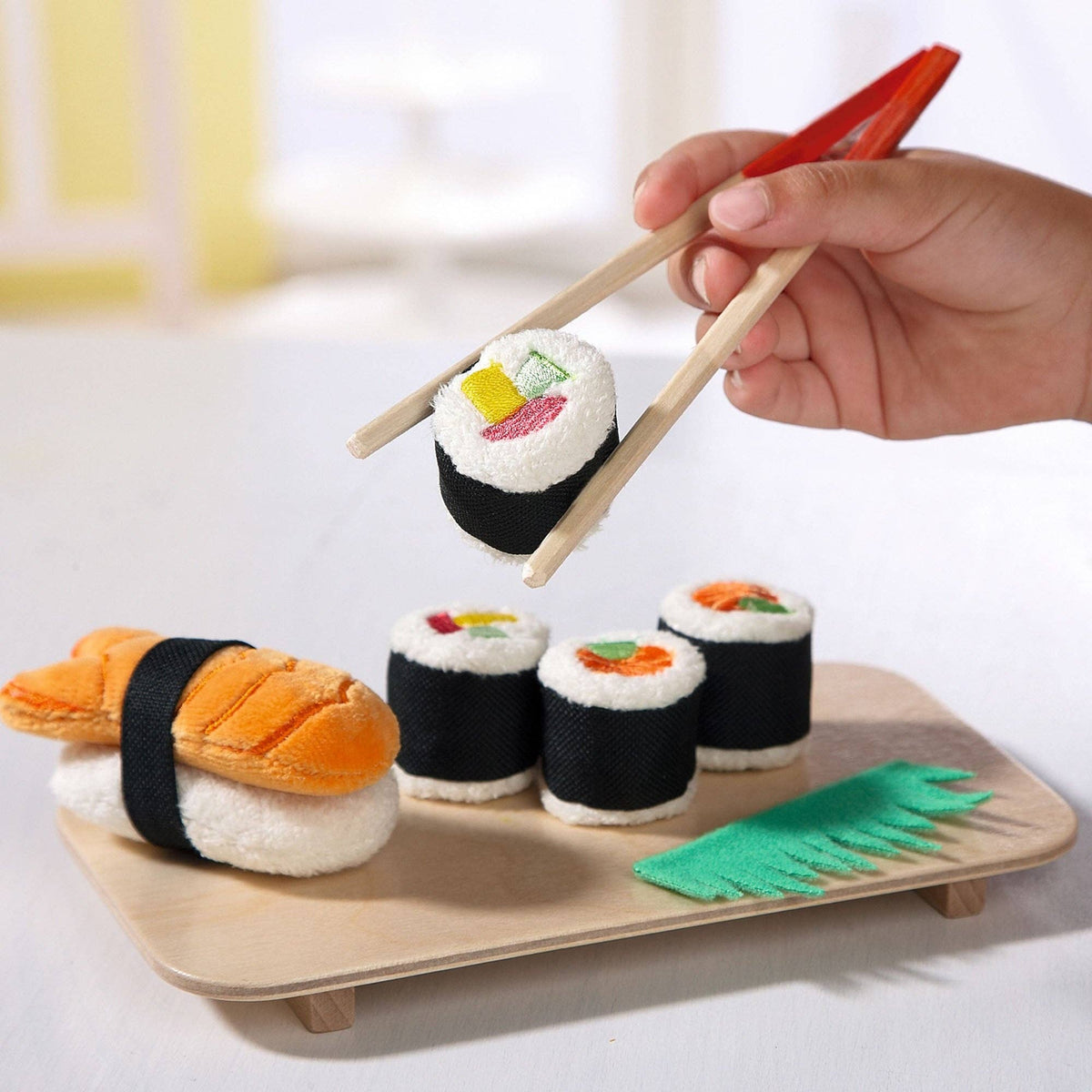 Hand holding chopsticks over a set of plush sushi toys on a wooden tray.