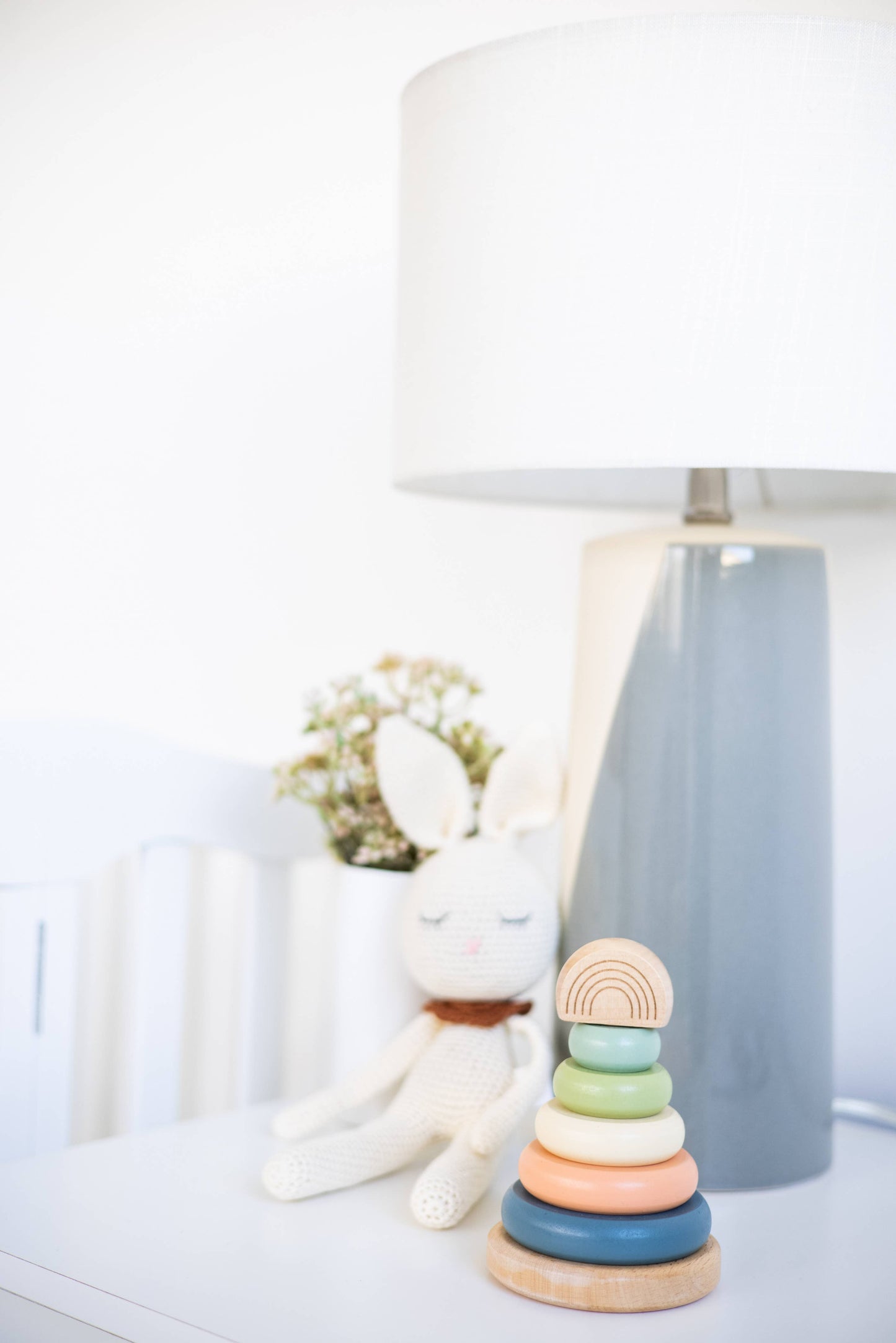 A wooden stacking toy consisting of a peg with five multicolor earth tone rings and a wooden rainbow topper sitting on table next to a white bunny toy and blue lamp