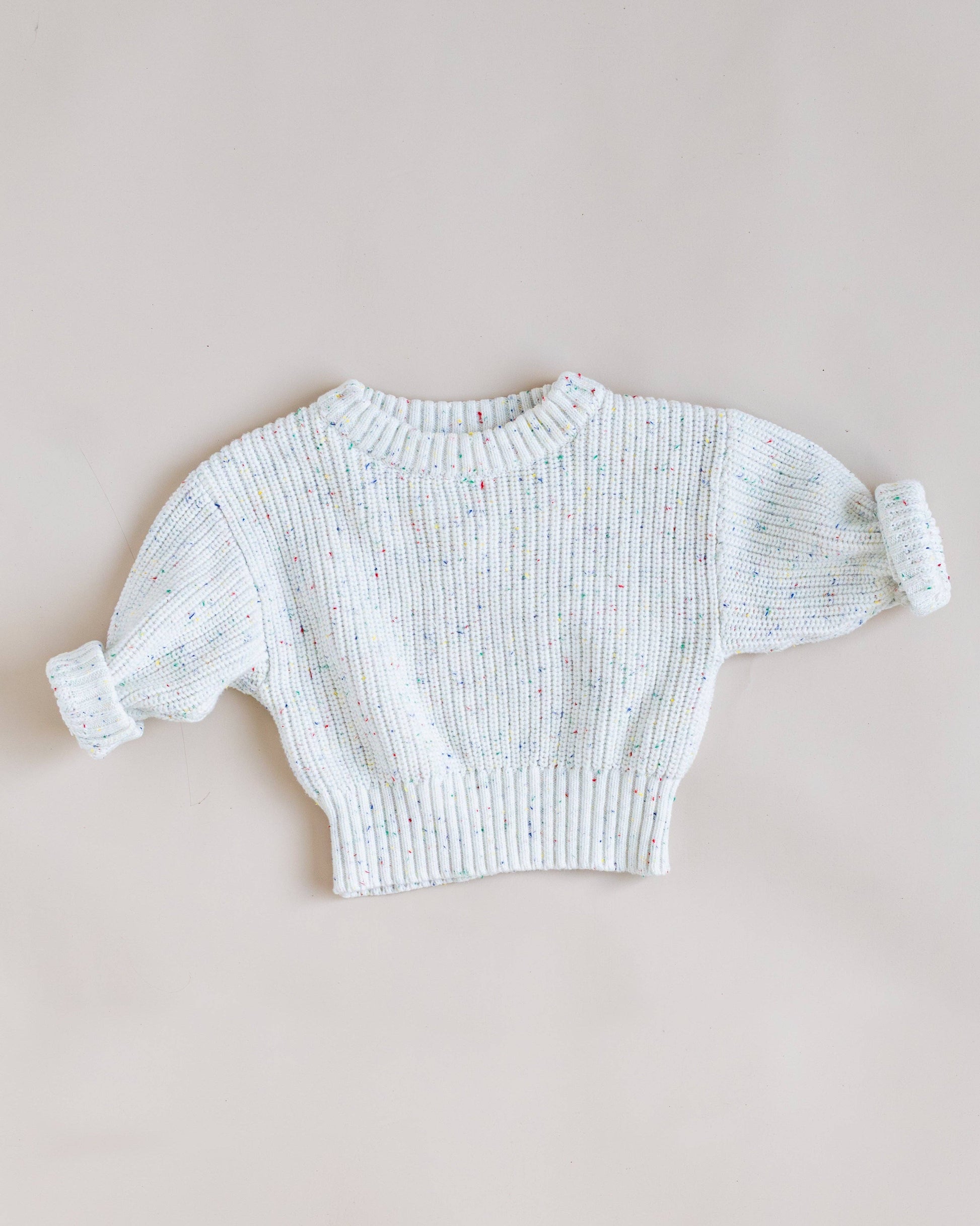 A white ribbed knit sweater with multicolored flecks, featuring voluminous short sleeves with rolled cuffs, laid flat on a neutral beige background.