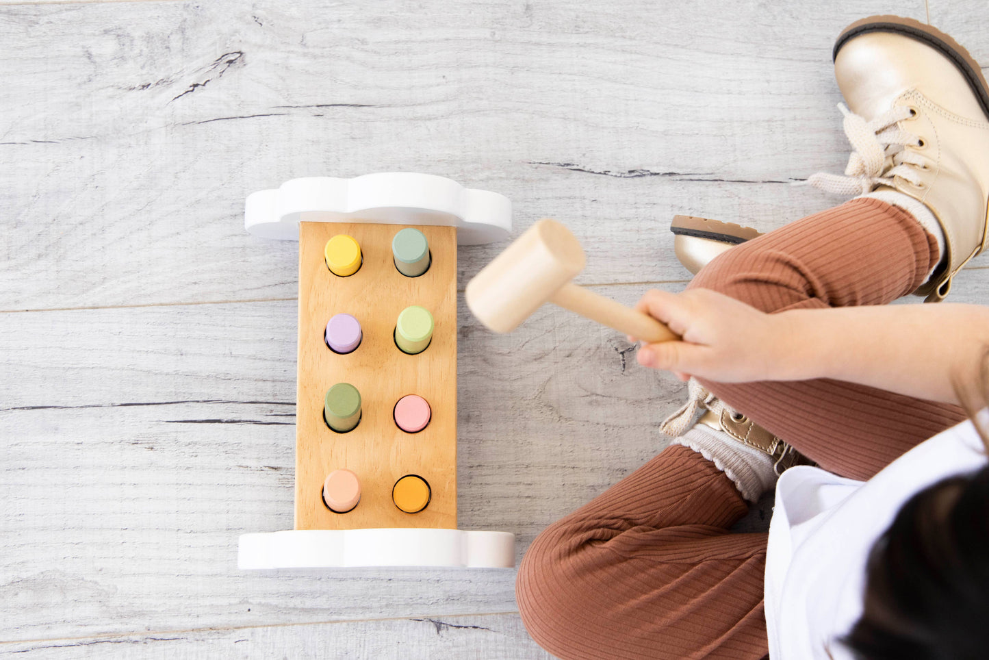 an aerial view of a young child sitting on a wooden floor playing with a wooden hammer bench toy with colorful pegs and a wooden mallet.