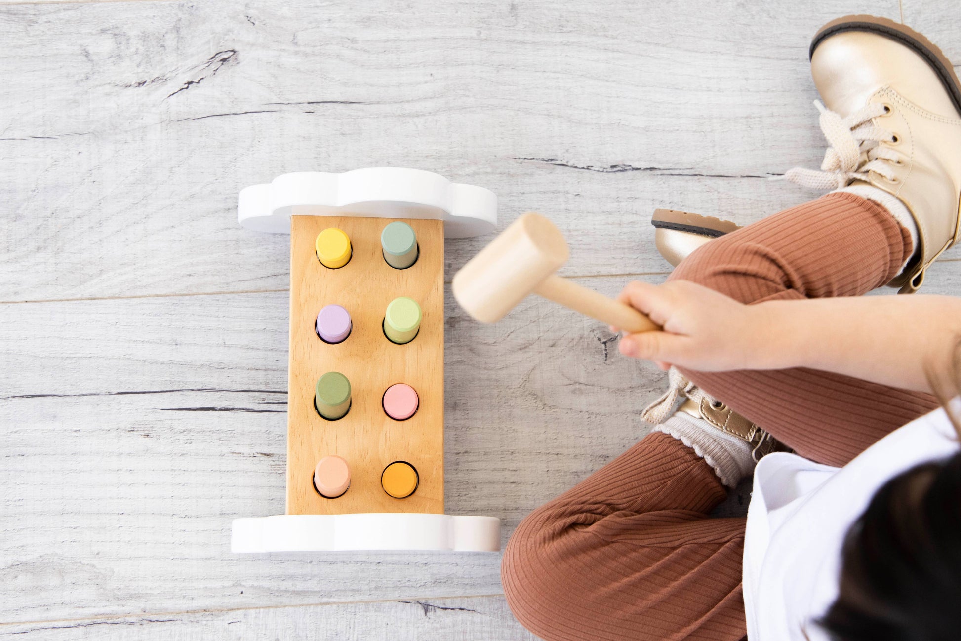 an aerial view of a young child sitting on a wooden floor playing with a wooden hammer bench toy with colorful pegs and a wooden mallet.