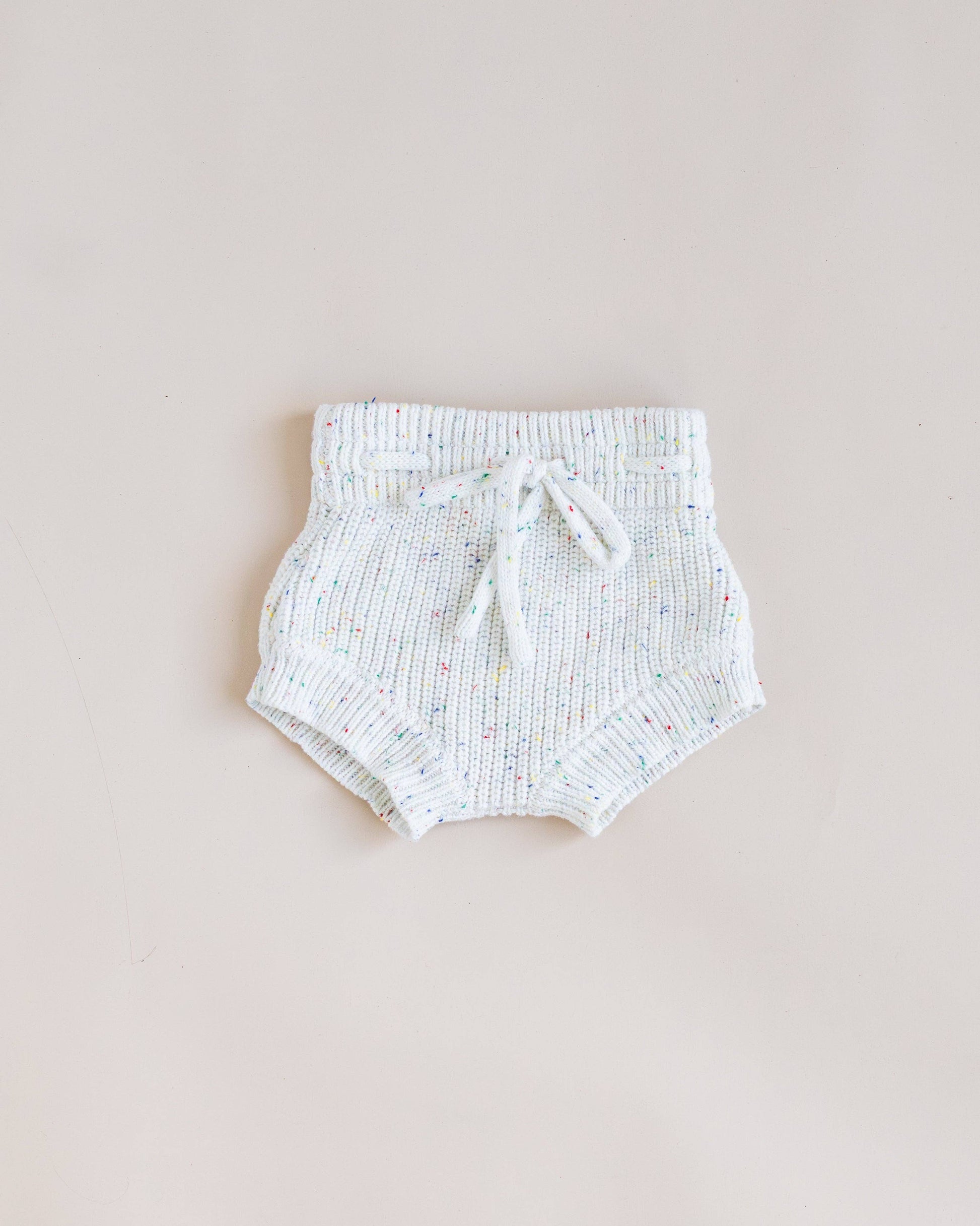 A pair of white knit baby bloomers with a textured ribbed pattern and a matching drawstring tie, laid flat on a light beige background.