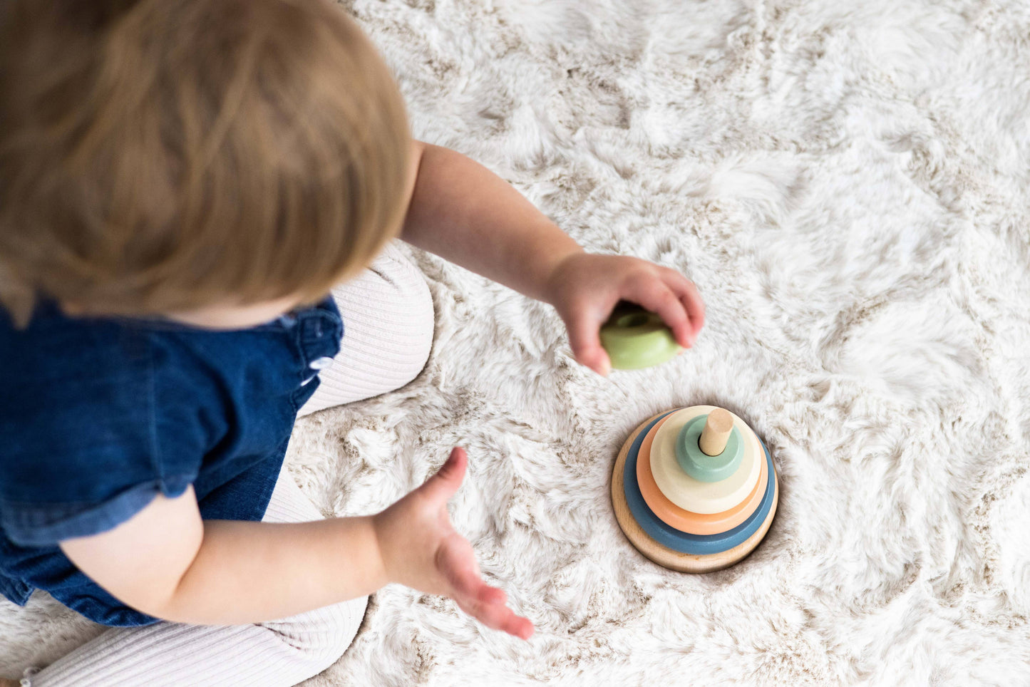 An aerial view of a child sitting on a fluffy carpet playing with a wooden stacking toy consisting of a peg with five multicolor earth tone rings.