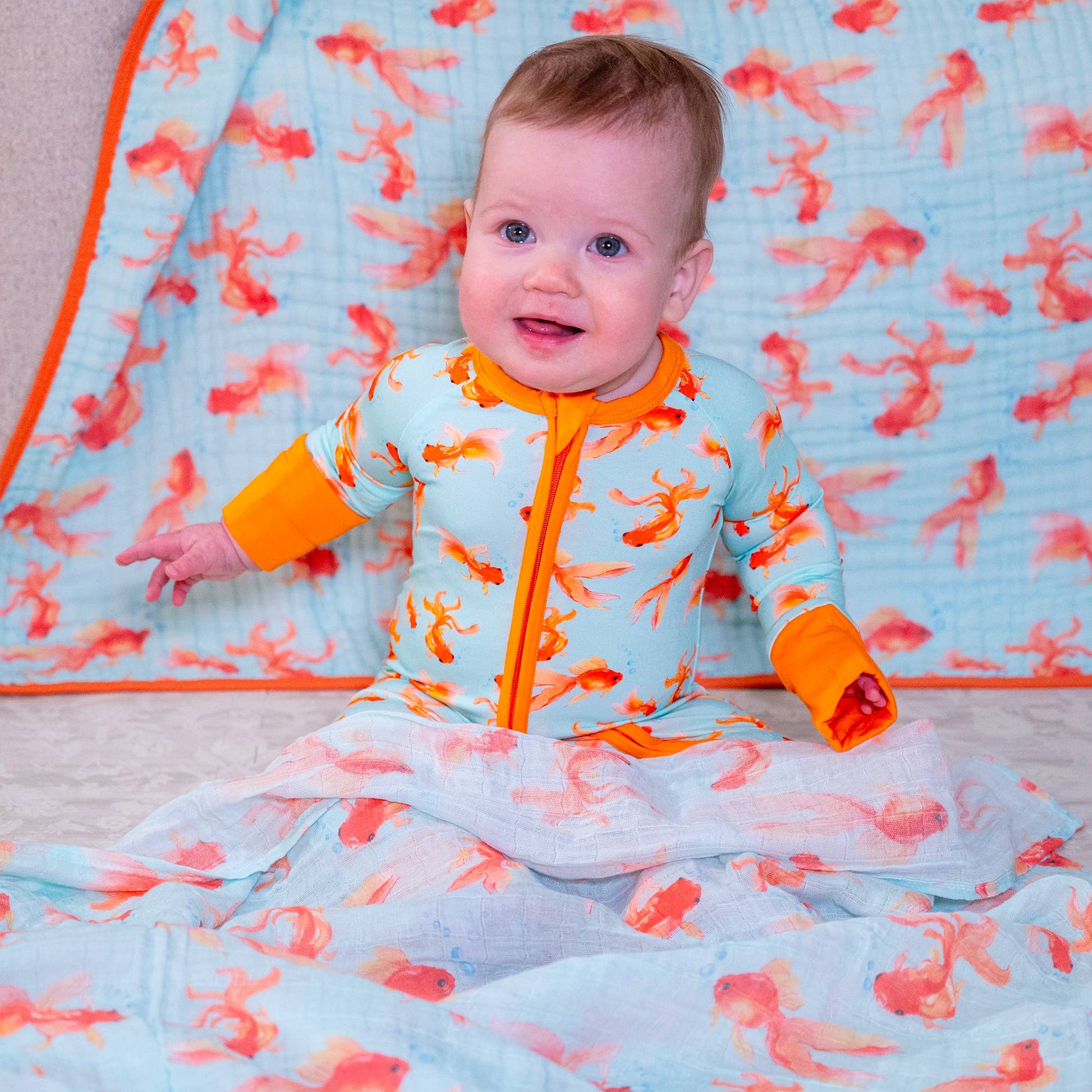 Baby in an orange and blue onesie pajama with goldfish pattern on a matching blanket.