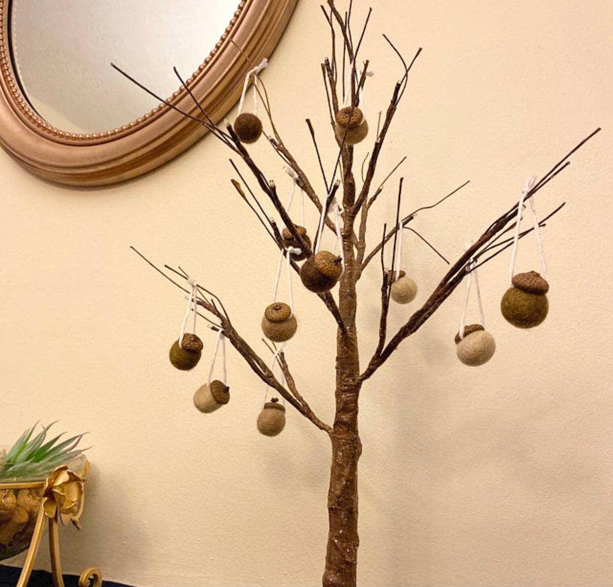 A decorative artificial tree branch display with multiple acorn ornaments hanging from the branches against a beige wall, with part of a round mirror and a small plant visible.