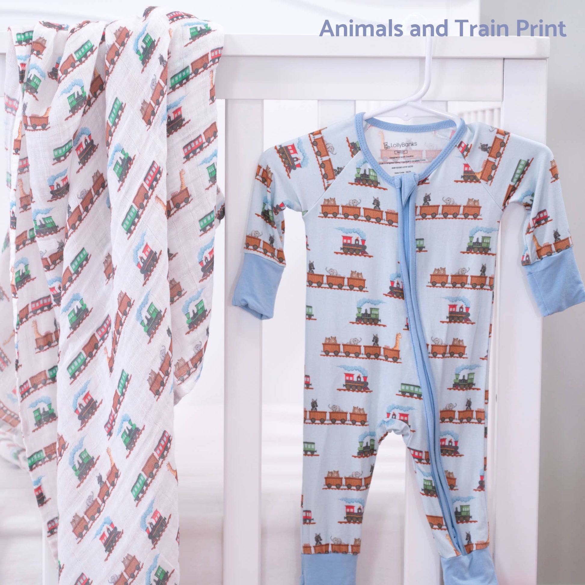 A baby footed one-piece sleeper and matching swaddle blanket, both featuring a repeating train print, displayed on a white crib. The sleeper is light blue with brown and green trains, and the swaddle is white with diagonal stripes of the same train pattern.