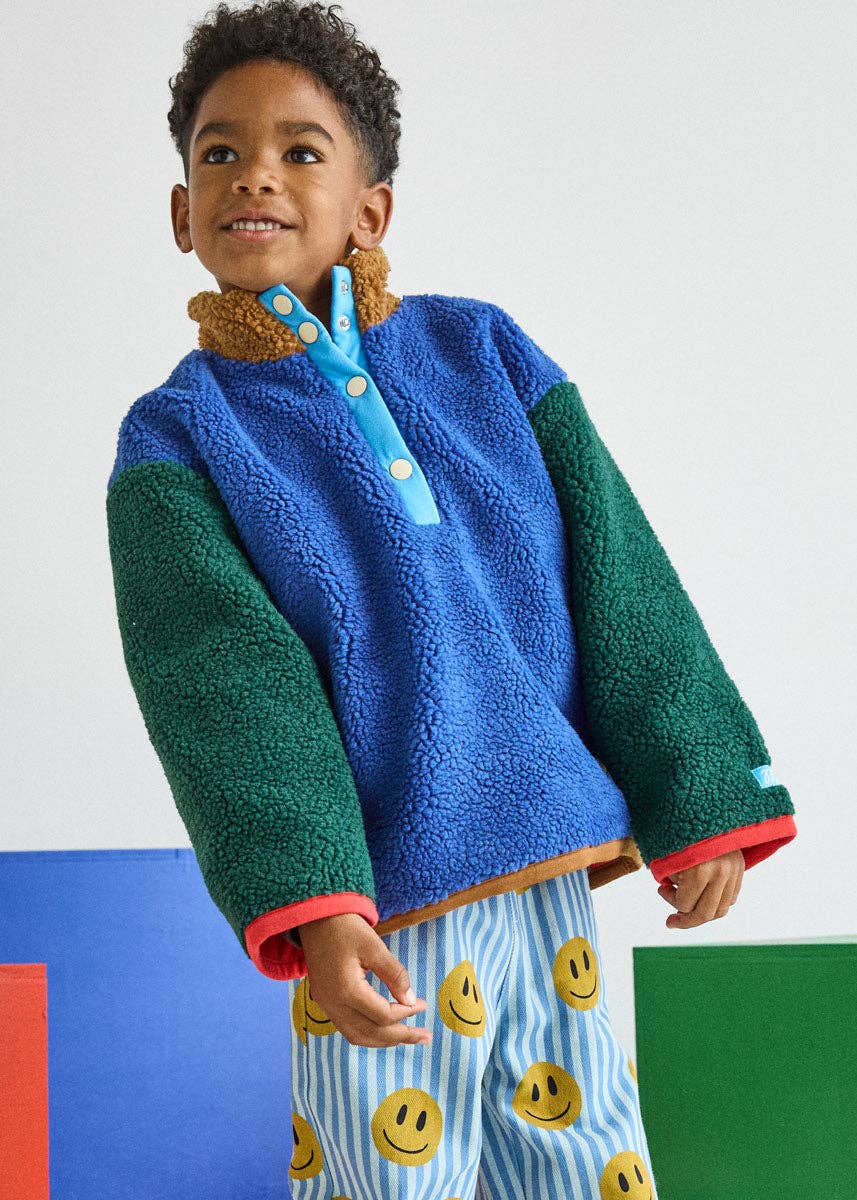 A young boy wearing a colorful fleece pullover with a blue body, green sleeves, and a brown teddy bear collar, paired with blue and white smiley face striped jeans, posing against a studio background.