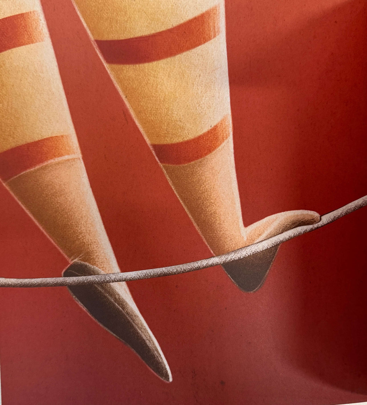 A close-up of a red and beige striped socked leg with a pointed toe, balanced on a thin wire against a solid red background.