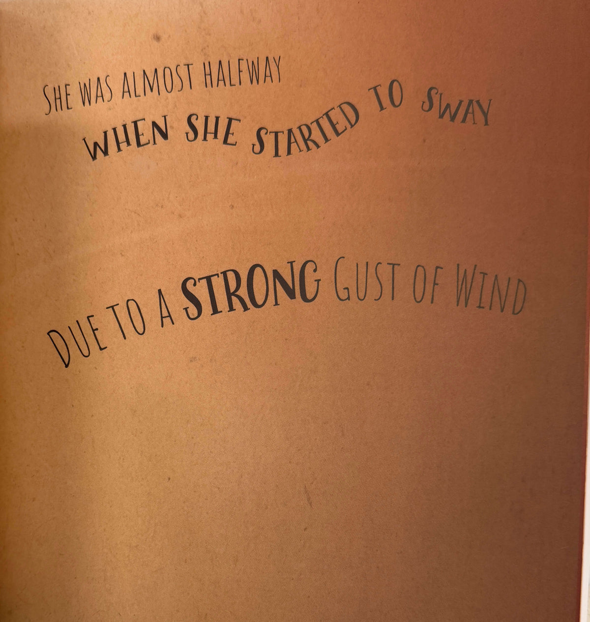 A close-up of a brown kraft paper surface with printed black text reading, 'SHE WAS ALMOST HALFWAY WHEN SHE STARTED TO SWAY DUE TO A STRONG GUST OF WIND.'.