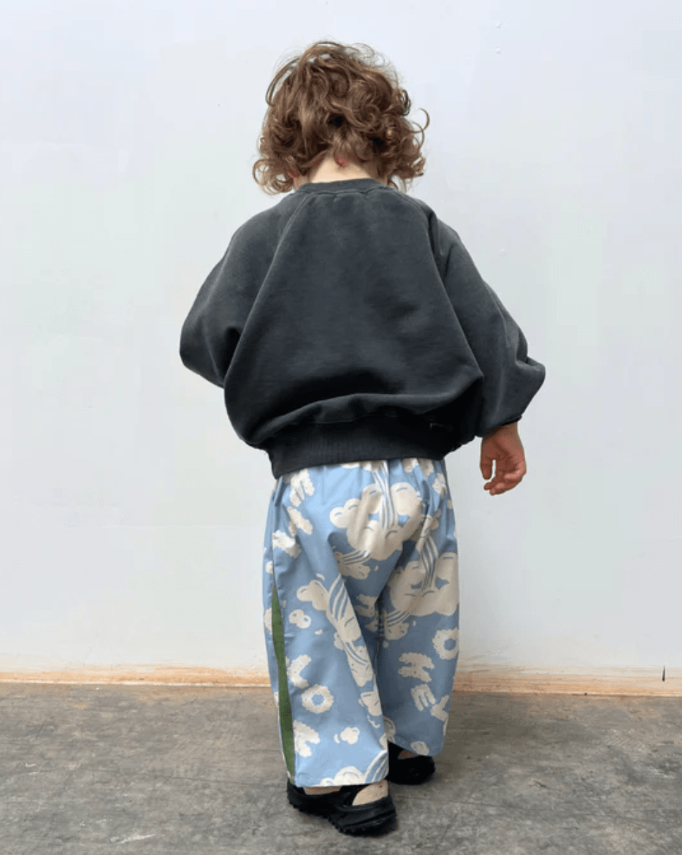 Child wearing a dark sweater and blue patterned pants standing against a white wall.