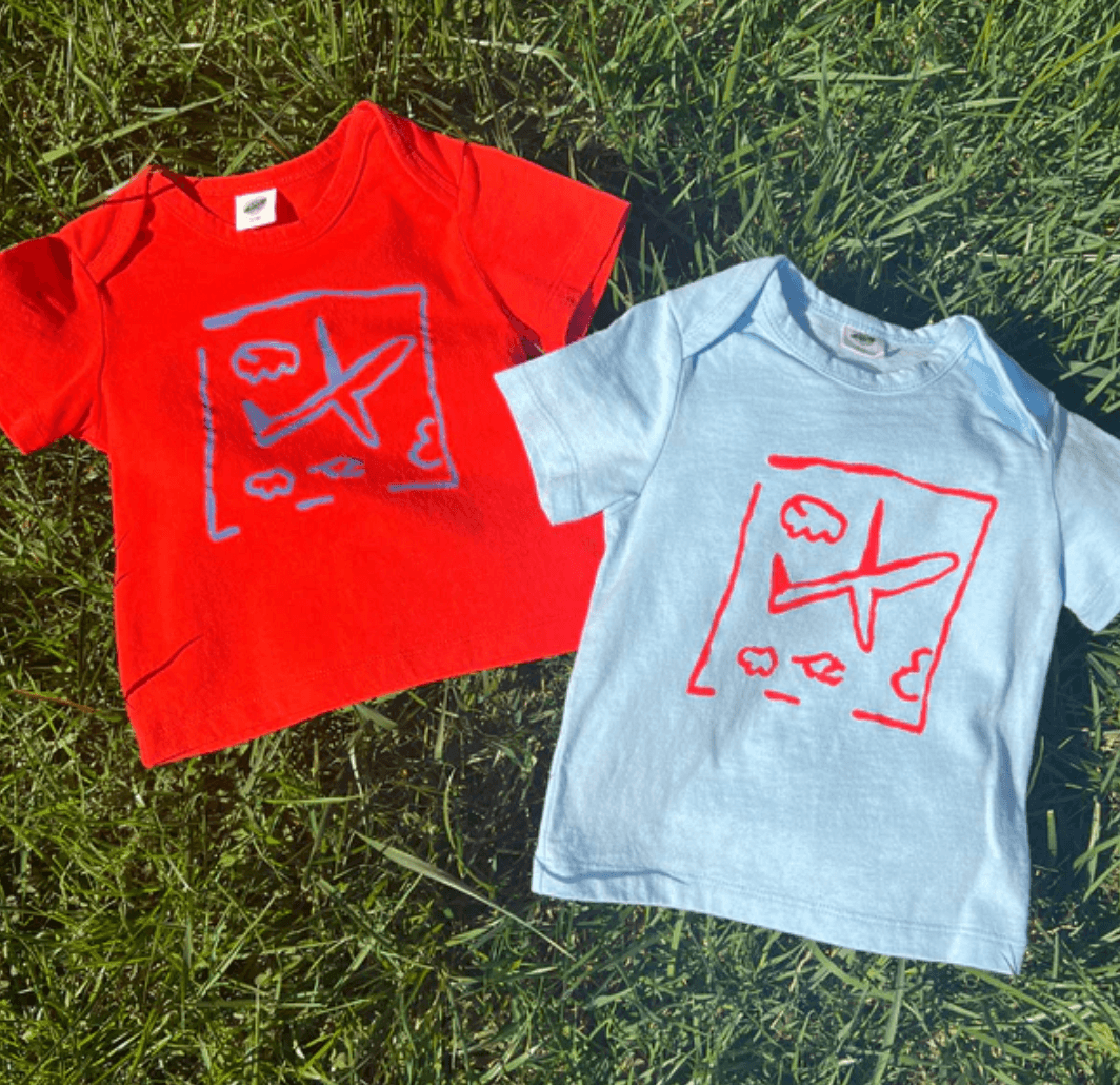 Two children's t-shirts, one red and one light blue, with graphic designs on a grass background.