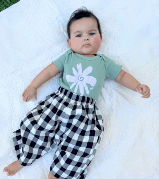 Baby lying on a white blanket wearing a green shirt with a flower design and black and white checkered pants.