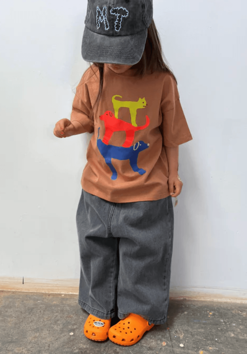 Child wearing a brown shirt with colorful dog design, gray cap, gray pants, and orange shoes against a white wall.