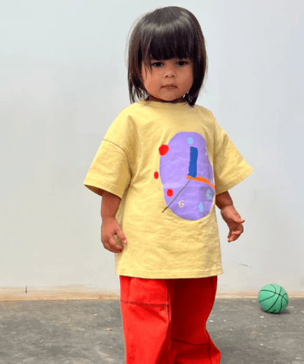 Child wearing a yellow shirt with a colorful design and red pants, standing on a gray floor with a white wall background.