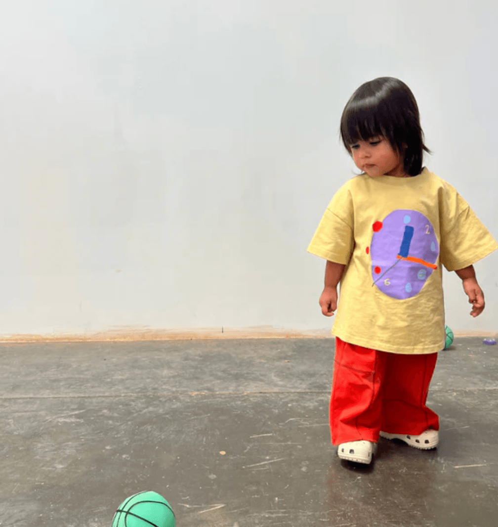 Child wearing a yellow shirt with a colorful design and red pants standing on a gray floor.