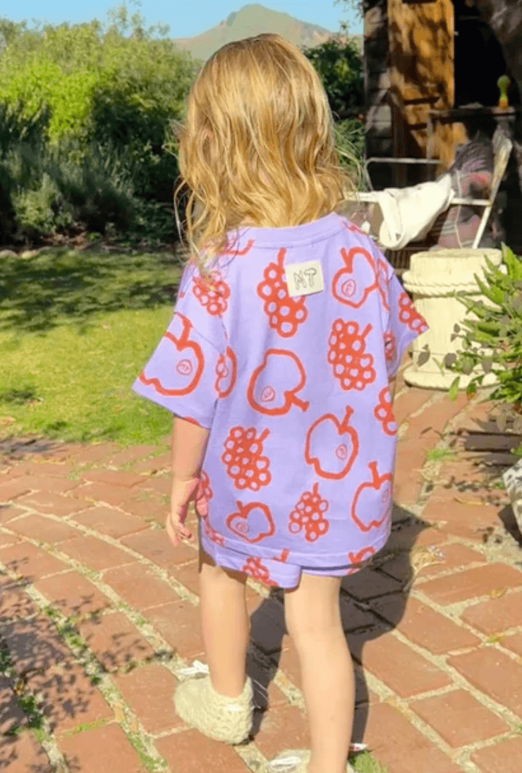 Child wearing a purple shirt with red fruit patterns outdoors on a brick path.