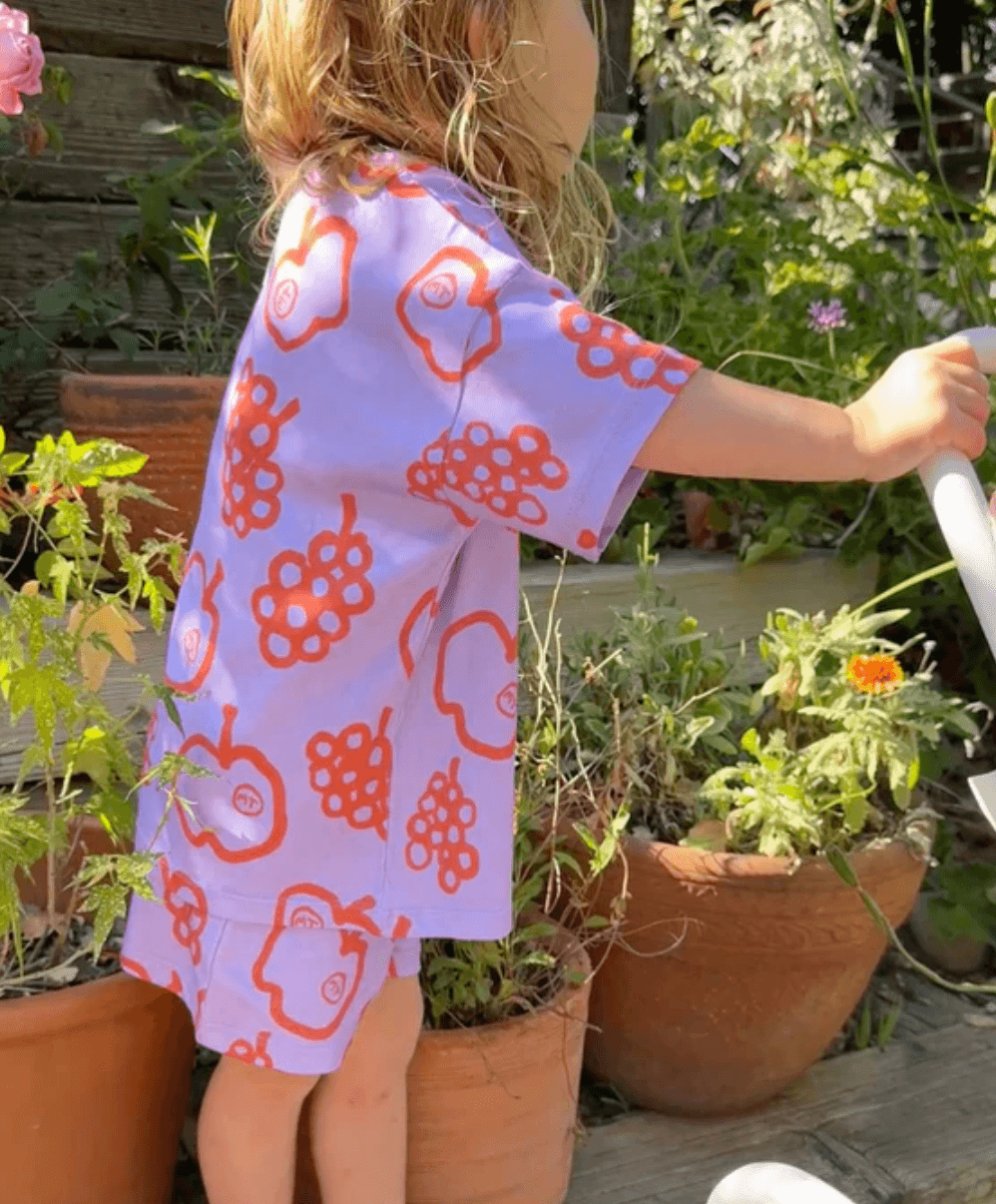 Child in a purple short set with red floral patterns watering plants in a garden.