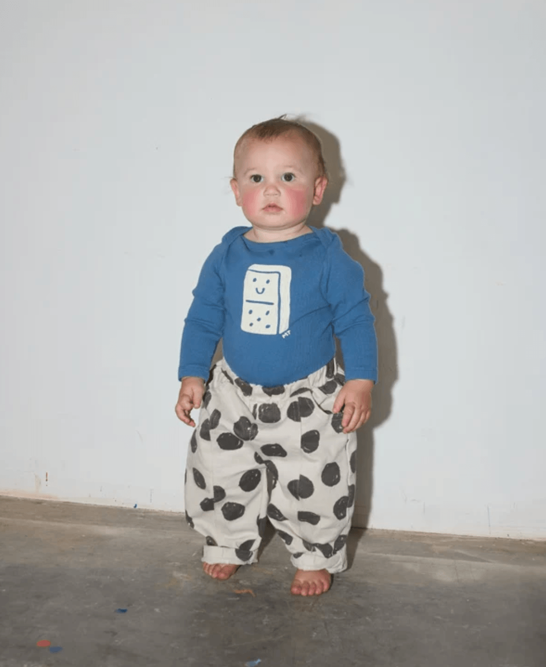 A baby standing barefoot against a plain white wall, wearing a blue long-sleeve top with a white domino graphic and loose white pants covered in black spots.