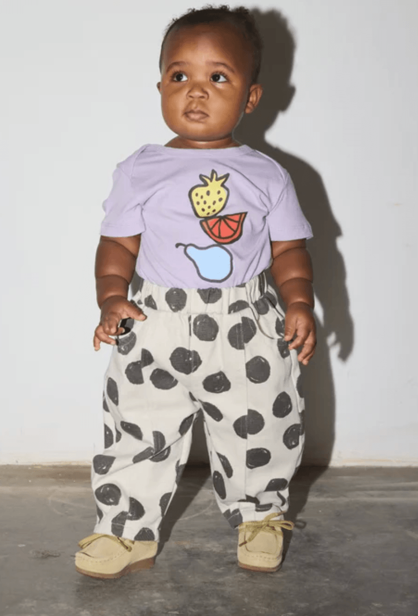 A baby standing against a plain wall wearing a light purple t-shirt with a fruit graphic and cream-colored pants with black spots, paired with tan shoes.