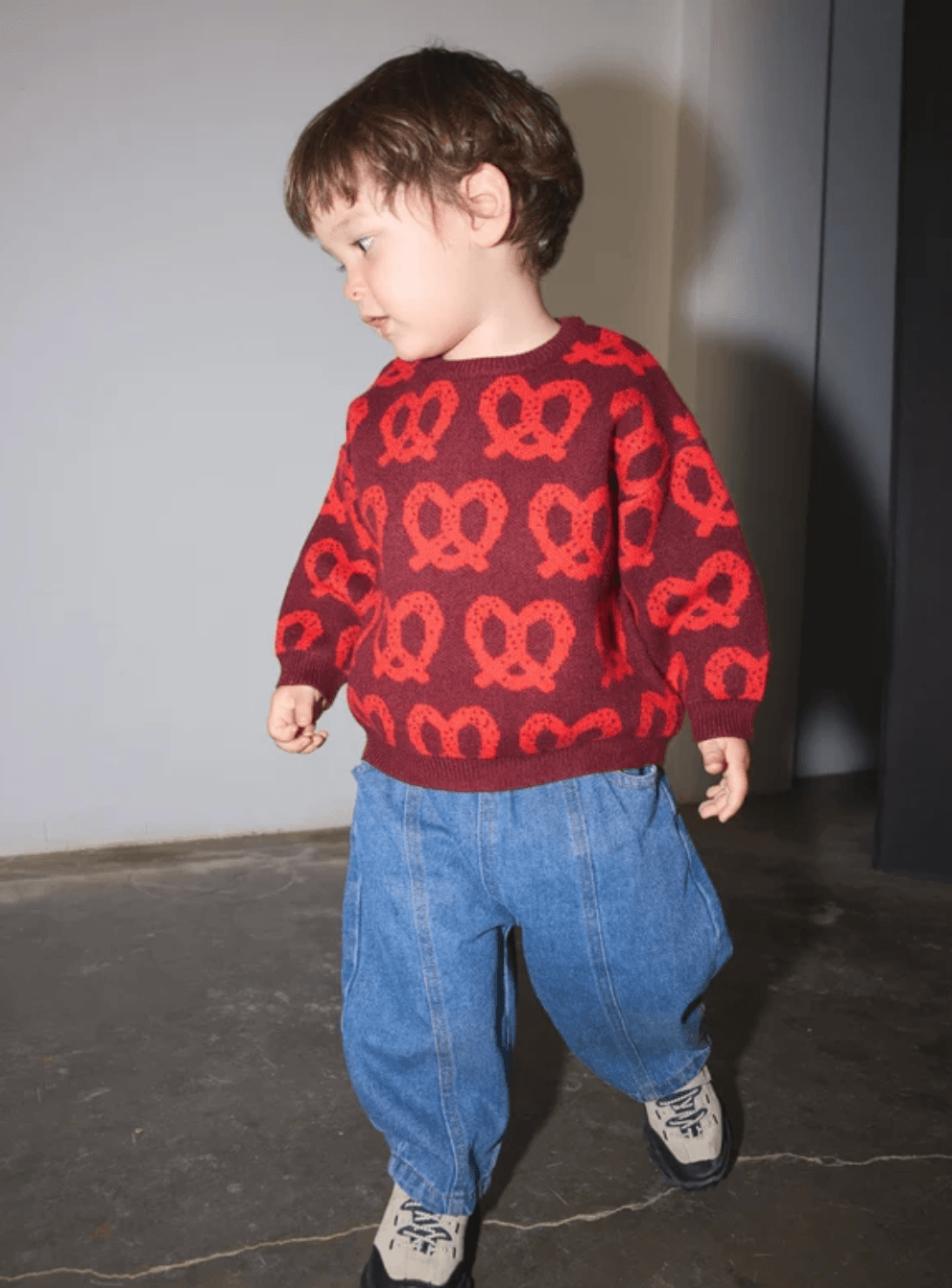 A toddler wearing a red knit sweater with a red pretzel pattern, paired with loose blue jeans and black-and-white sneakers, standing in an indoor setting.
