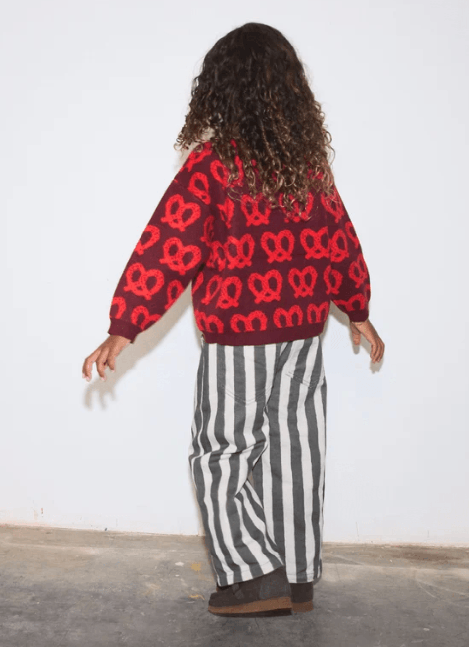 A child viewed from behind wearing a red knit sweater with a repeating red heart motif and wide-leg black-and-white vertically striped pants, standing against a plain light wall.