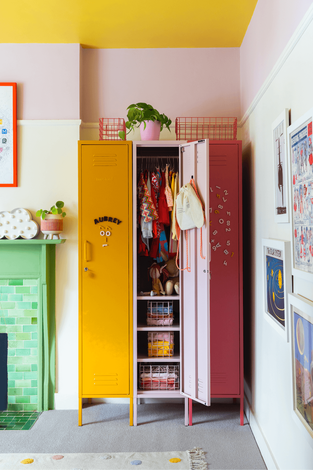 A set of three colorful children's lockers in yellow, pink, and red, styled in a playful room with a green tiled fireplace and framed art on the walls.