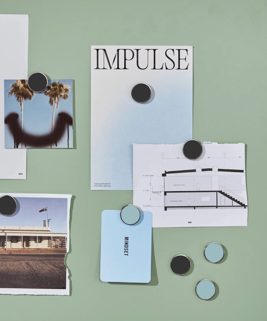 A magnetic memo board filled with printed cards, papers, and circular magnets on a pale green background. Visible printed pieces include one with the word IMPULSE, one with a palm tree photo, one with a building floor plan, and one light blue card with the word MINDSET.