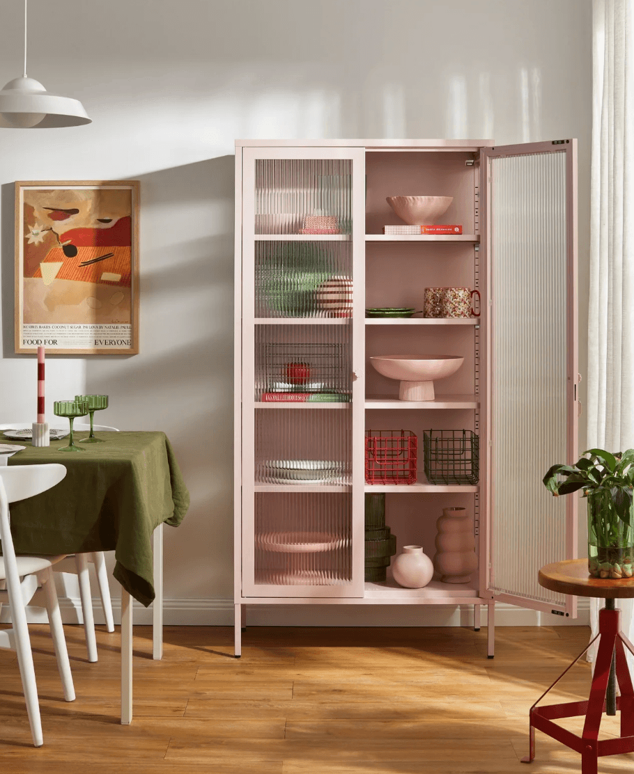 A tall, light pink metal cabinet with two frosted glass doors stands open in a styled dining room. The cabinet has multiple shelves holding pink and green dishes, bowls, and vases, set against a warm wood floor and a white wall with soft sunlight shadows.