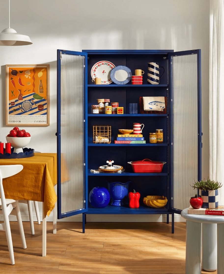 A tall navy blue open storage cabinet with glass doors stands in a bright kitchen or dining room, filled with dishes, books, and decorative items. The room has light wood floors, a white wall, and a table with a mustard yellow cloth on the left.