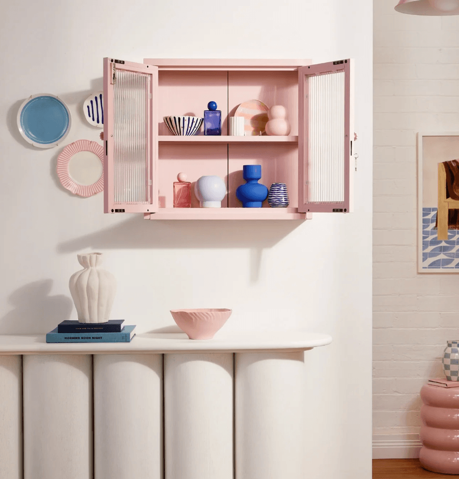 A pink wall-mounted cabinet with open doors and wire-frame glass doors, styled with pastel dishes and vases inside, set against a white wall above a white console table.