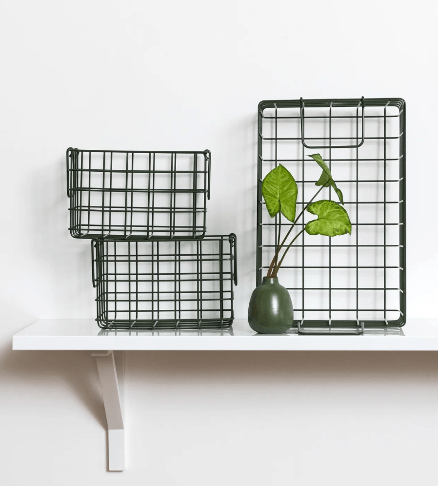 Three dark green wire grid baskets in different sizes arranged on a white floating shelf, with a small green vase holding green leaves placed in front of the largest basket.