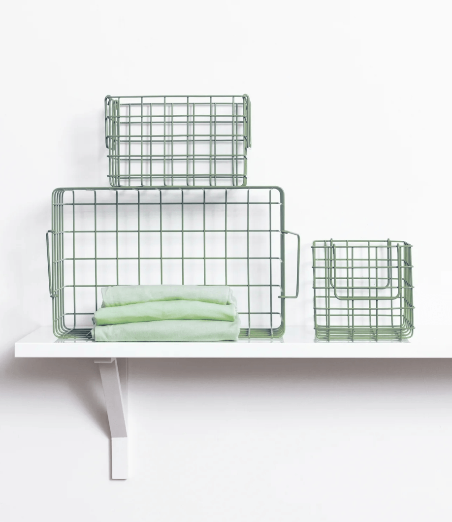 Three light green wire grid storage baskets of different sizes, with a folded pale green towel in the largest basket, displayed on a white wall-mounted shelf against a white background.