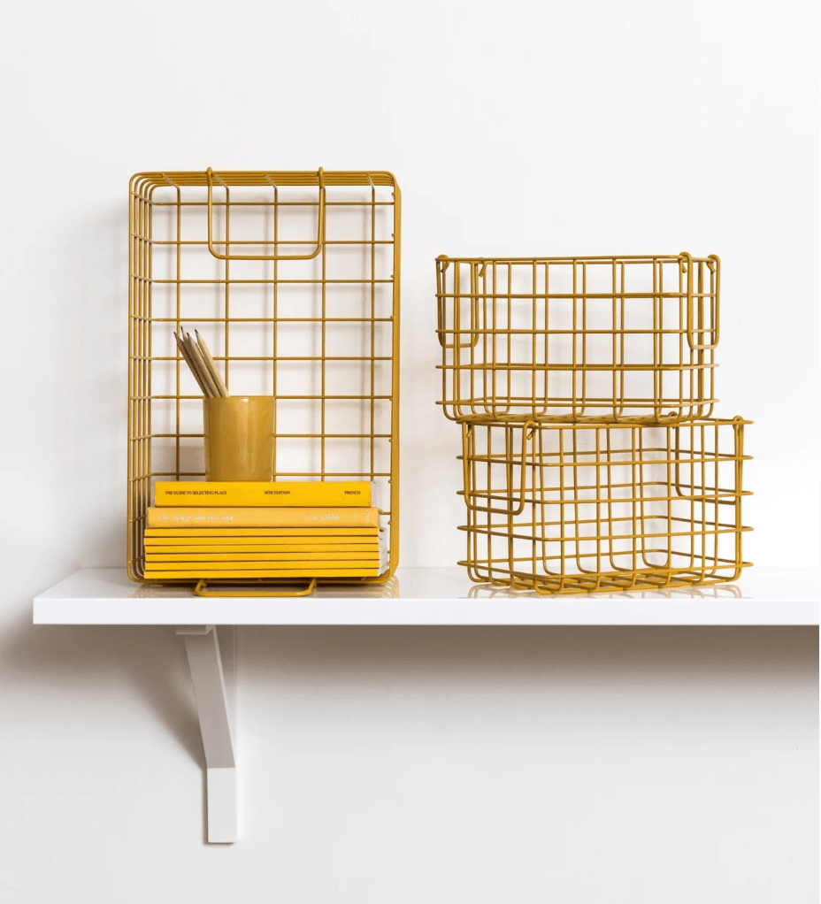 Three mustard yellow wire grid storage baskets displayed on a white shelf against a white wall, with one basket holding a yellow cup and books.