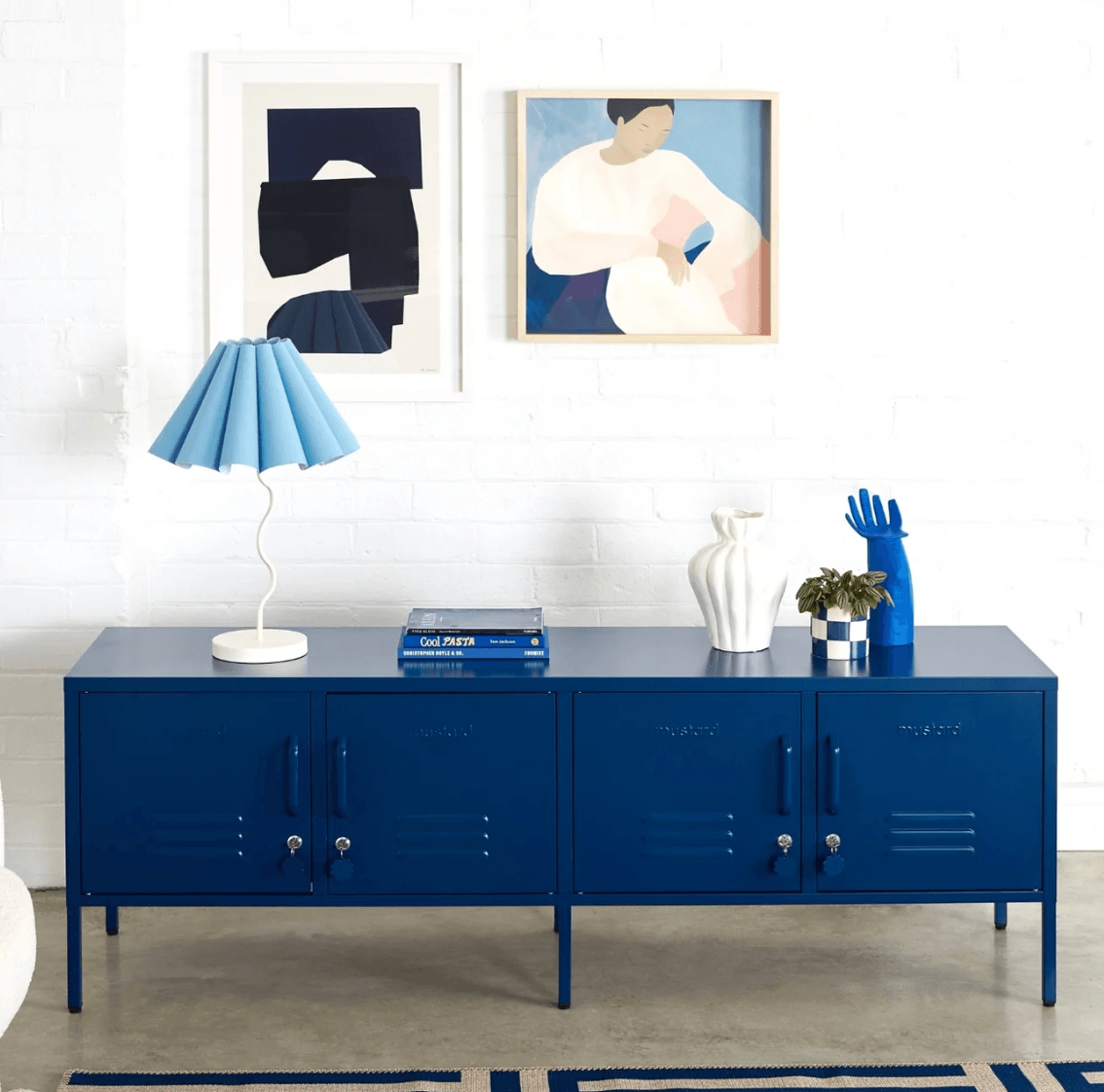 A long blue locker-style sideboard or media console with four cabinet doors, shown in a styled room setting with a blue pleated table lamp, white vases, a small plant, and framed wall art against a white brick wall.