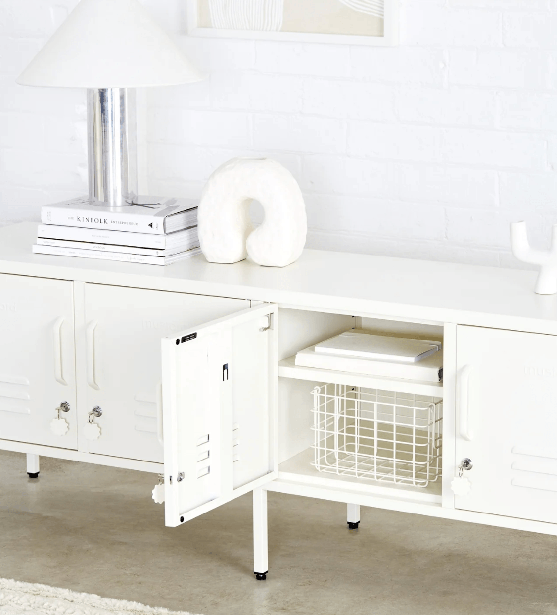 A white metal media console cabinet with two doors, one open to show storage space with a wire basket and books, set against a white brick wall with a lamp and decorative items on top.