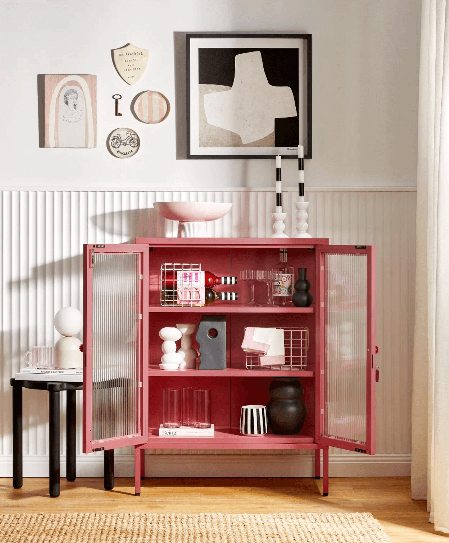 A pink metal cabinet with two ribbed glass doors open revealing three interior shelves and a variety of decor items arranged on the shelves.