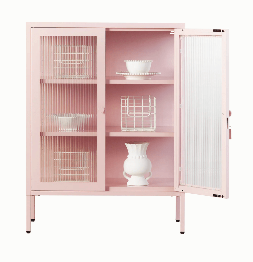 A light pink metal cabinet with two ribbed glass doors and three interior shelves, shown on a plain white background. One door is open revealing a light pink metal interior with white dishe sand baskets arranged on shelves.