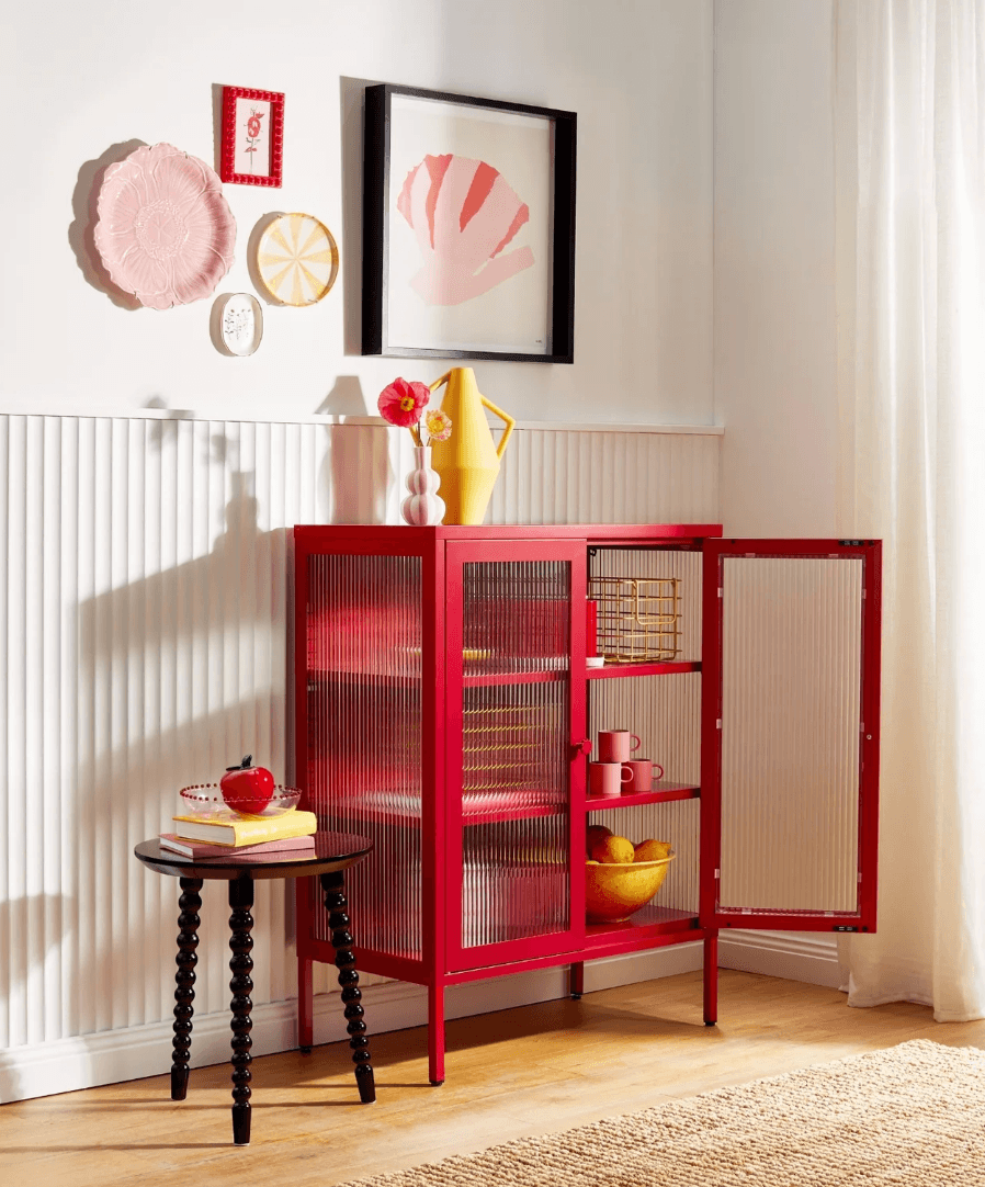 A red metal  storage cabinet with glass doors and panels stands in a bright room with a black side table, yellow vase, and framed wall art.